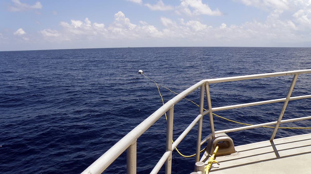 Looking over the bow railing at the lines attaching a boat to a mooring buoy in the water