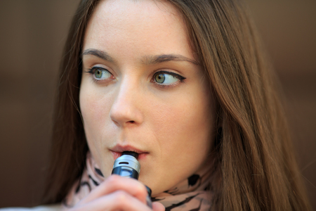 Vape teenager. Young pretty white girl in casual clothing smoking an electronic cigarette opposite modern brown background on the street in the spring. Bad habit. Vaping activity. Close up.