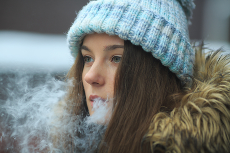 Vape teenager. Young pretty white girl in blue cap is smoking an electronic cigarette on the street in the winter. Bad habit. Close up.