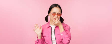 Stinky. Asian girl in sunglasses and stylish outfit, shuts her nose from disgust, dislike bad smell, standing over pink background