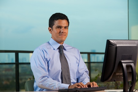 Successful hispanic office worker using a computer in an office with great view. 30s latino American male of mixed Brazilian - Mexican descent.