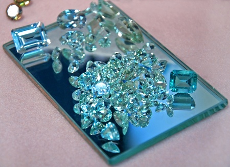 This stock image shows loose aquamarine gemstones in both cut rectangular shapes and round diamond cut shapes on a rectangle mirror, full of sparkle and shine.