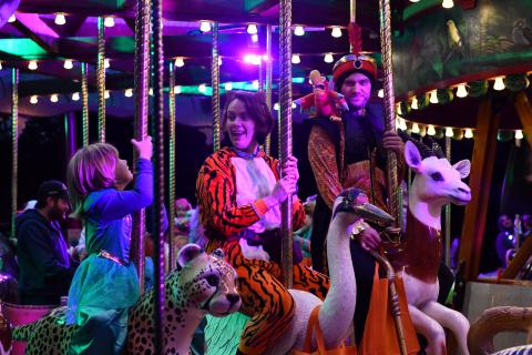 Photo of a family of two adults and a young child riding a carousel together. All three people are wearing Halloween costumes.