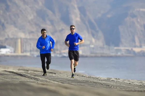 Two men friends running together on beach sand with beautiful coast mountain background in morning training session jogging workout one in long sleeve and pants the other guy in shorts