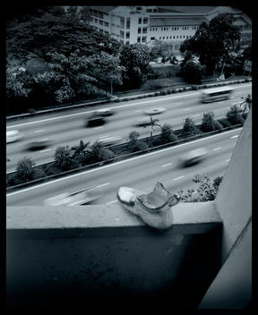 A Monopoly Icon Shoe On The Edge Of A Building Wall Looking Down On Heavy Traffic Highway.