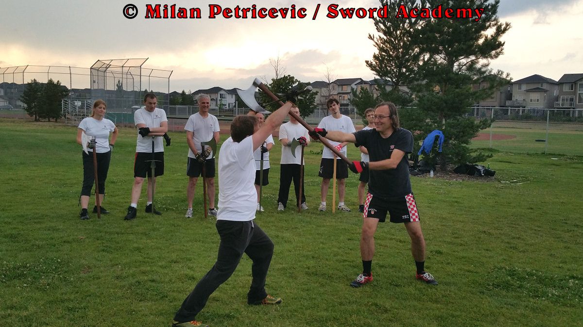 Milan demonstrates during an outdoor class in front of Sword Academy students two handed axe against the armored sword exercise overpowering the opponent with the momentum of the two handed axe as inspired by historical sources from the German medieval (and renaissance) tradition, part of Sword Academy HEMA / WMA / Martial Arts curriculum.