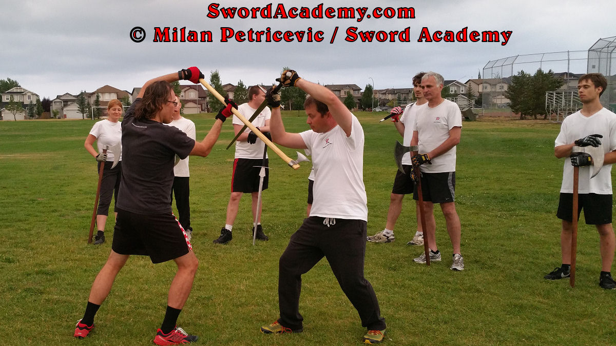 Milan demonstrates during an outdoor class in front of Sword Academy students two handed axe against the armored sword exercise switching attacks using the nimble two handed axe as inspired by historical sources from the German medieval (and renaissance) tradition, part of Sword Academy HEMA / WMA / Martial Arts curriculum.