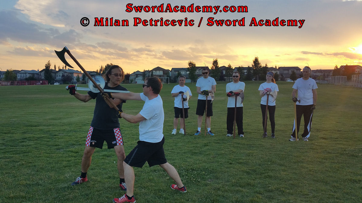 Milan demonstrates during an outdoor class in front of Sword Academy students defending with sword and buckler against the two handed axe attack exercise counter-striking and thrusting with the sword, while maintaining pressure with buckler as inspired by historical sources from the German medieval (and renaissance) tradition, part of Sword Academy HEMA / WMA / Martial Arts curriculum.