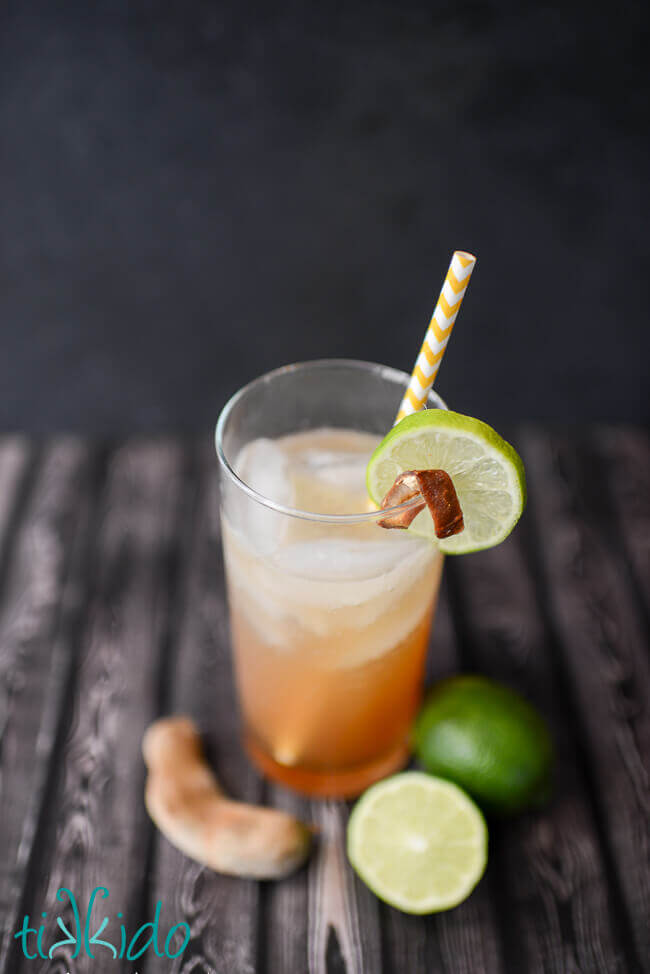 Tamarind margarita in a tall glass with a yellow and white striped paper straw, slice of lime garnish, and peel of tamarind garnish.  Limes and tamarind pods on wooden surface around glass.