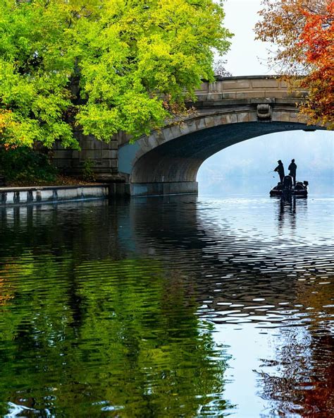 two people on a small boat in the water under an overpass with fall foliage