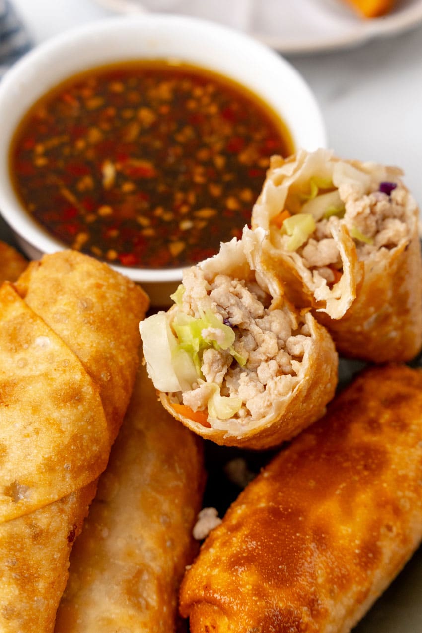 a homemade egg roll broken in half and resting on a plate of egg rolls with a small white bowl filled with sesame garlic dipping sauce on the side