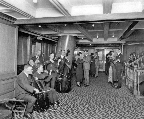 Musicians play in the C-Deck Second Class Entrance foyer of the Aft Second Class Staircase aboard the RMS Olympic.
The doors to the far left granted access to the Second Class Library, and the large slanted tube in the center of the room is the base...