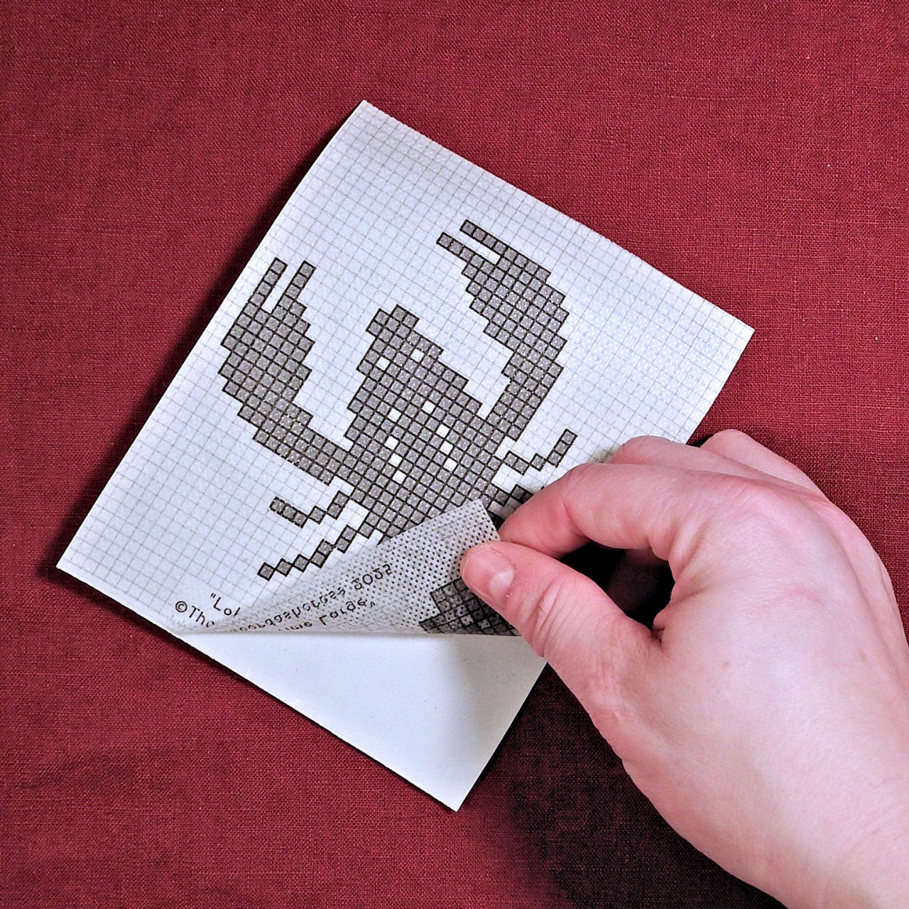 Photograph of the lobster embroidery patch, with a hand lifting up the corner to show it peels off like a sticker.