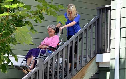 Senior in a wheelchair goes down the stairs using Stair-Trac with the help of a lady in a blue shir