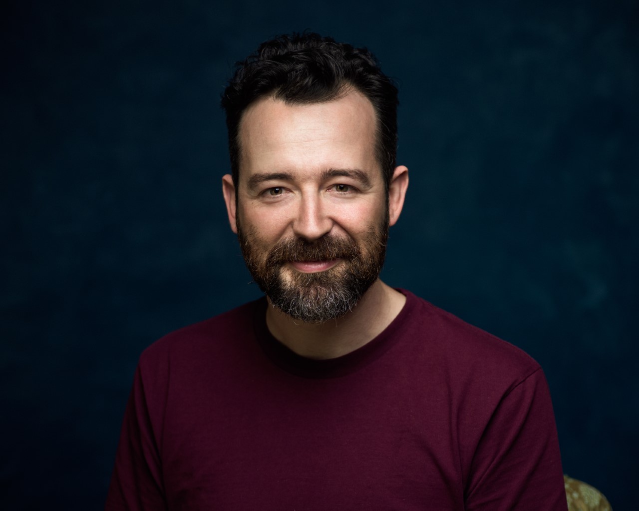 A headshot of Sam Smith, who has dark hair and a beard, and a slight smile as he looks down the barrel of the camera