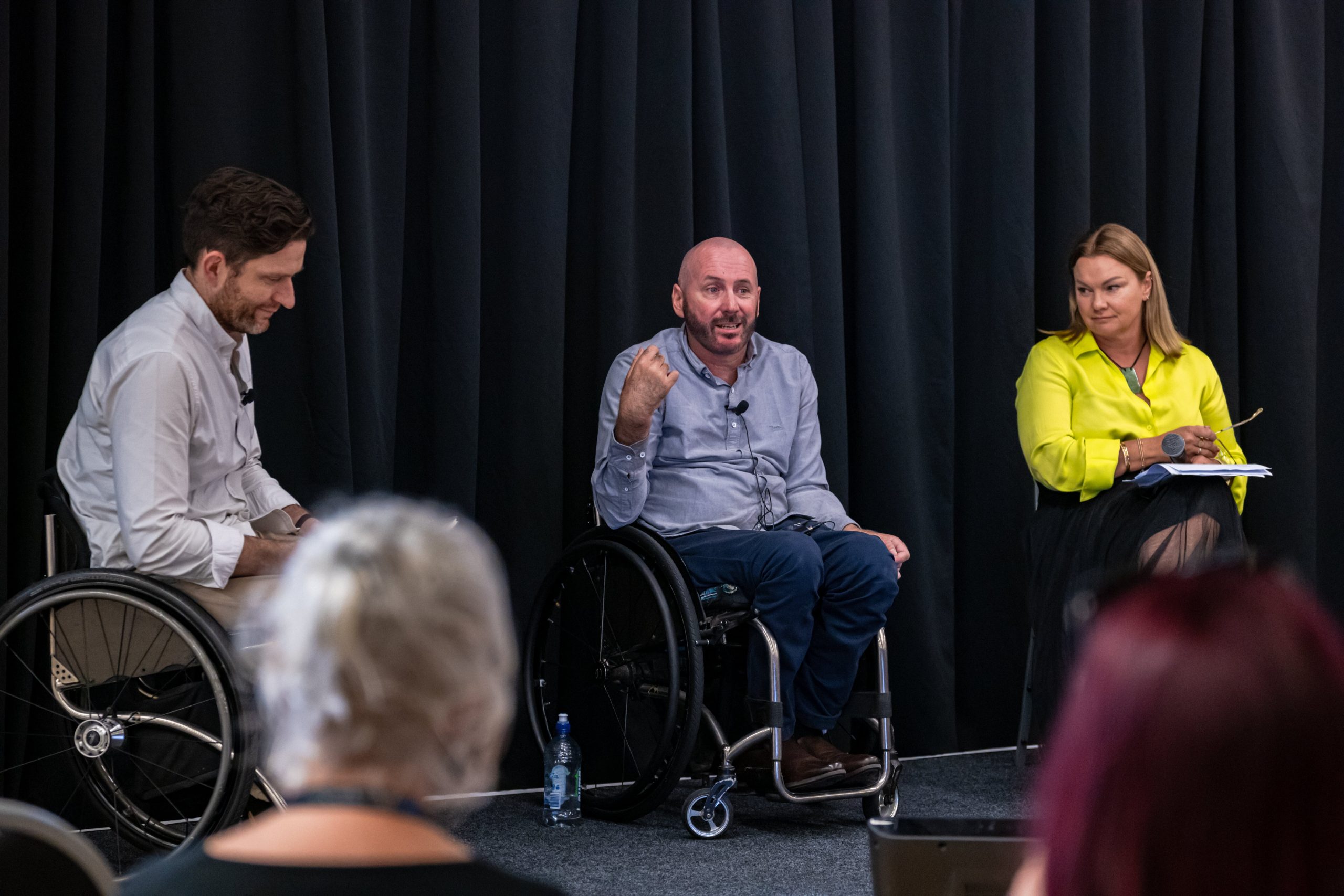 Dan Buckingham, Jai Waite and Rachale Davis are on a stage together. Dan has brown hair, a white button-up shirt and is using a wheelchair, turned away from the camera. Jai is wearing a blue button-up top, using a wheelchair, and is in the middle of speaking. Rachale has blonde shoulder-length hair and a yellow top, looking over at Jai.