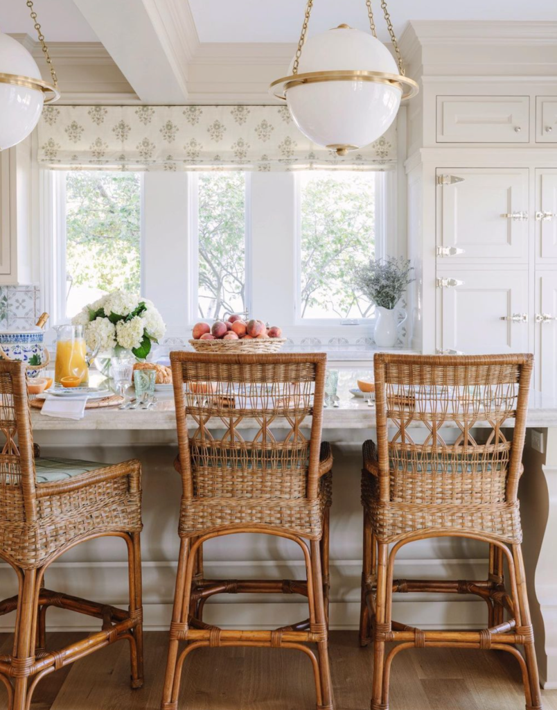 The kitchen features white cabinets, a soft blue and white tile backsplash, and a marble countertop. The barstools have a natural rattan finish and a cushioned seat, adding warmth and texture to the room. The kitchen has large windows that provide natural light. Decorative items such as a woven basket filled with peaches, a vase of flowers, and a wooden cutting board add to the coastal vibe of the space.