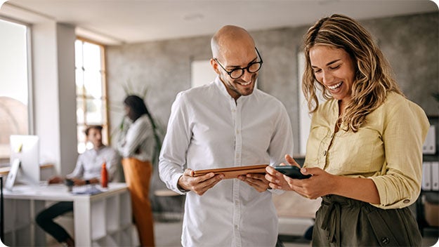 Four people in a room, tow in the background working at a Desktop computer, two in the front smiling, one holding a cell phone the other holding a tablet.