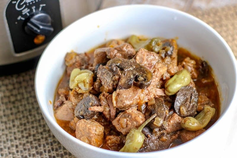 A white bowl filled with beef tips and pepperoncinis in front of crockpot.