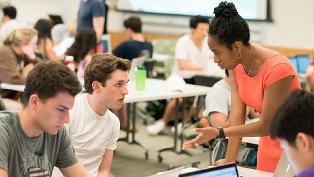 A teacher stands at a table next to two students and is speaking to one of them