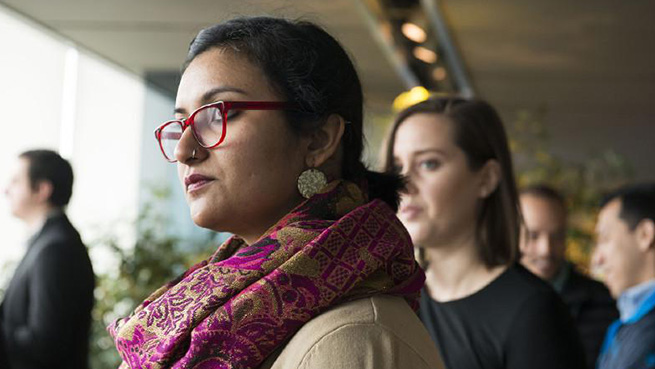 A group of people standing together out of focus. One woman is in front of the group, in focus but looking away from the camera. 