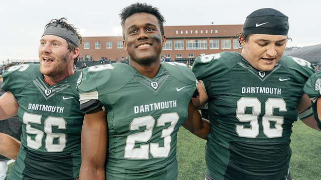 Three football student-athletes standing arm-in-arm on the football field during a Homecoming game.