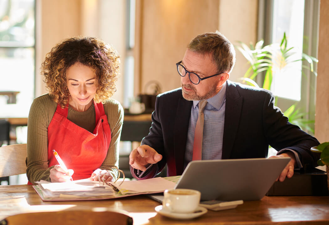 Woman in an apron writing in a binder and a man in a suit sitting next to her