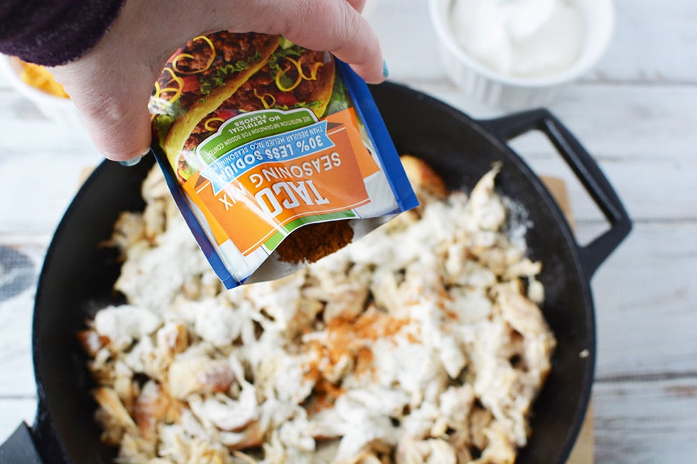 Pouring taco seasoning onto shredded chicken in a skillet. 