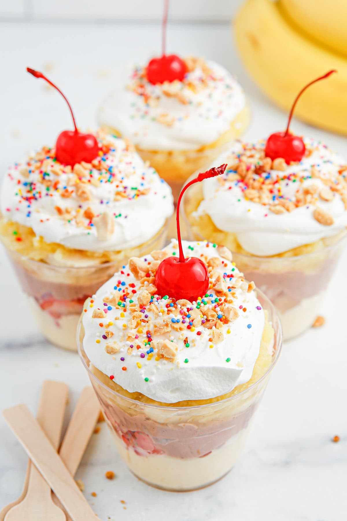 Four banana split pudding cups with whipped cream, sprinkles, and cherries.