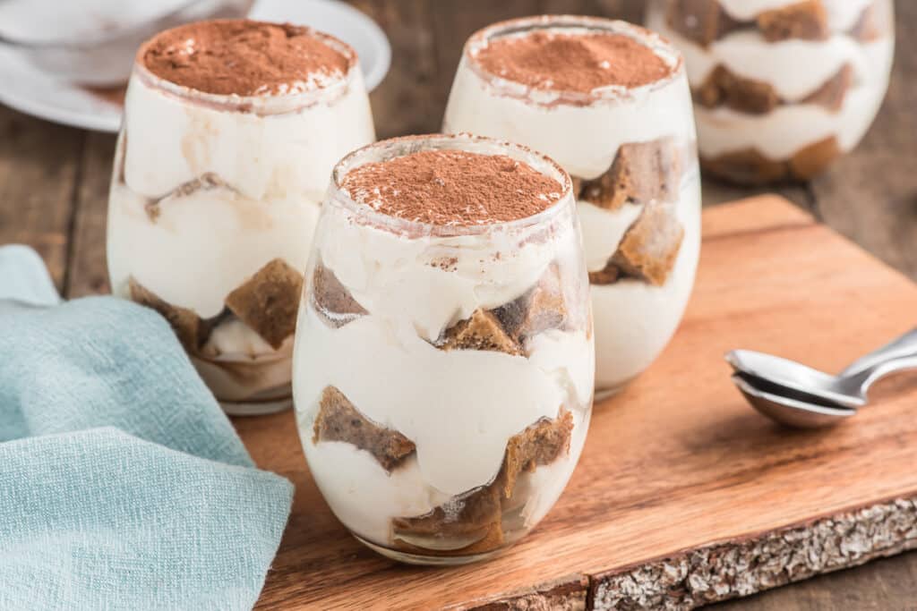 Three tiramisu parfaits in glasses on a wooden board.