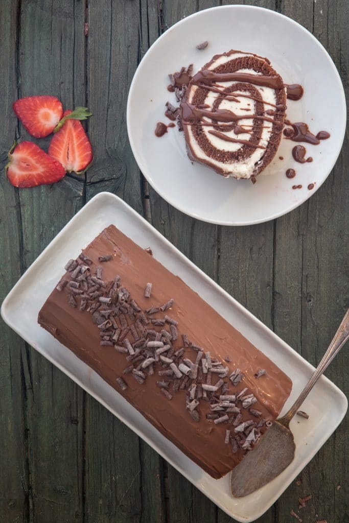 Roll cake on a white plate with a piece on a white plate drizzled with melted chocolate.