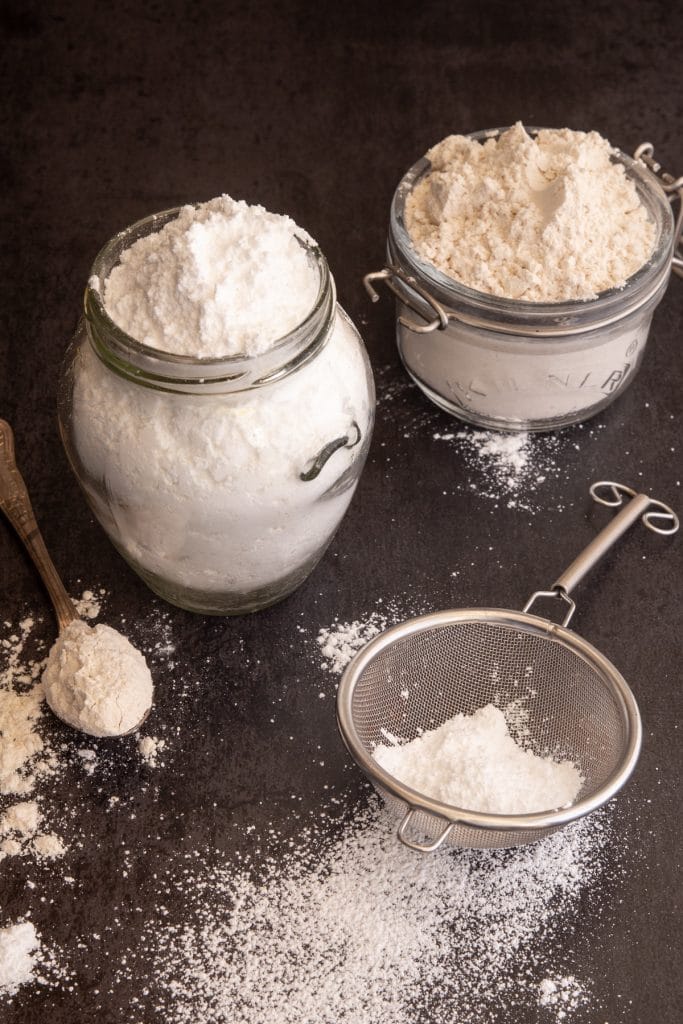 Brown and white powdered sugar in glass jars with some on a spoon and some in a whisk.