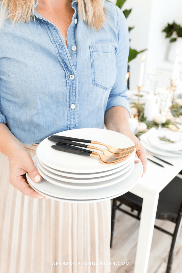 A person in a blue chambray shirt and beige skirt holds a stack of white dishes with gold and black utensils on top, ready for Thanksgiving dinner. The background shows a dining table set with plates, candles, and greenery. A website, apersonalorganizer.com, is visible at the bottom.