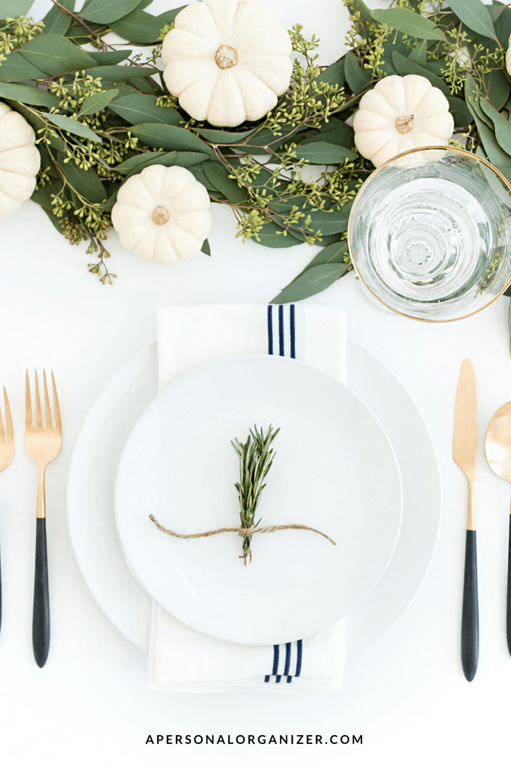 A white table is set with a plate, knife, fork, spoon, and glass. The plate has a small bundle of rosemary tied with twine on a white napkin with blue stripes. White pumpkins and greenery decorate the table, creating an inclusive space perfect for those with special needs. The URL "apersonalorganizer.com" is visible at the bottom.