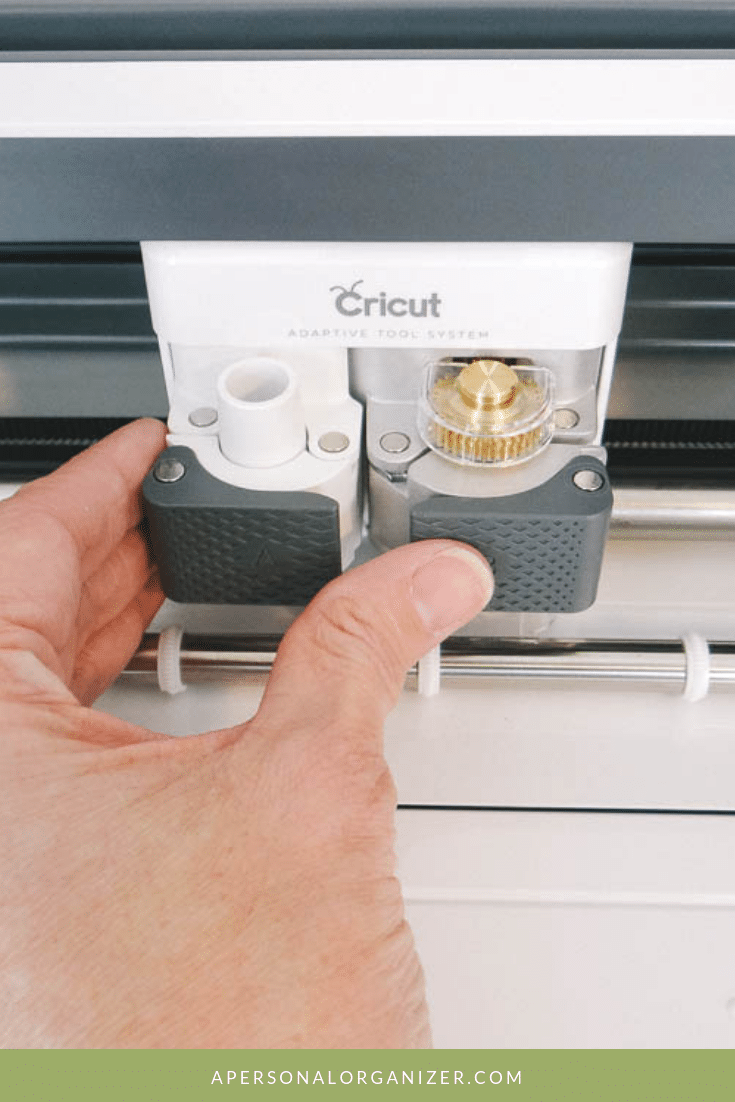 A close-up of a hand adjusting the tools in a Cricut Maker's adaptive tool system. The logo "Cricut" is visible on the machine. The environment looks clean and organized. At the bottom, a green strip displays the text "APERSONALORGANIZER.COM.