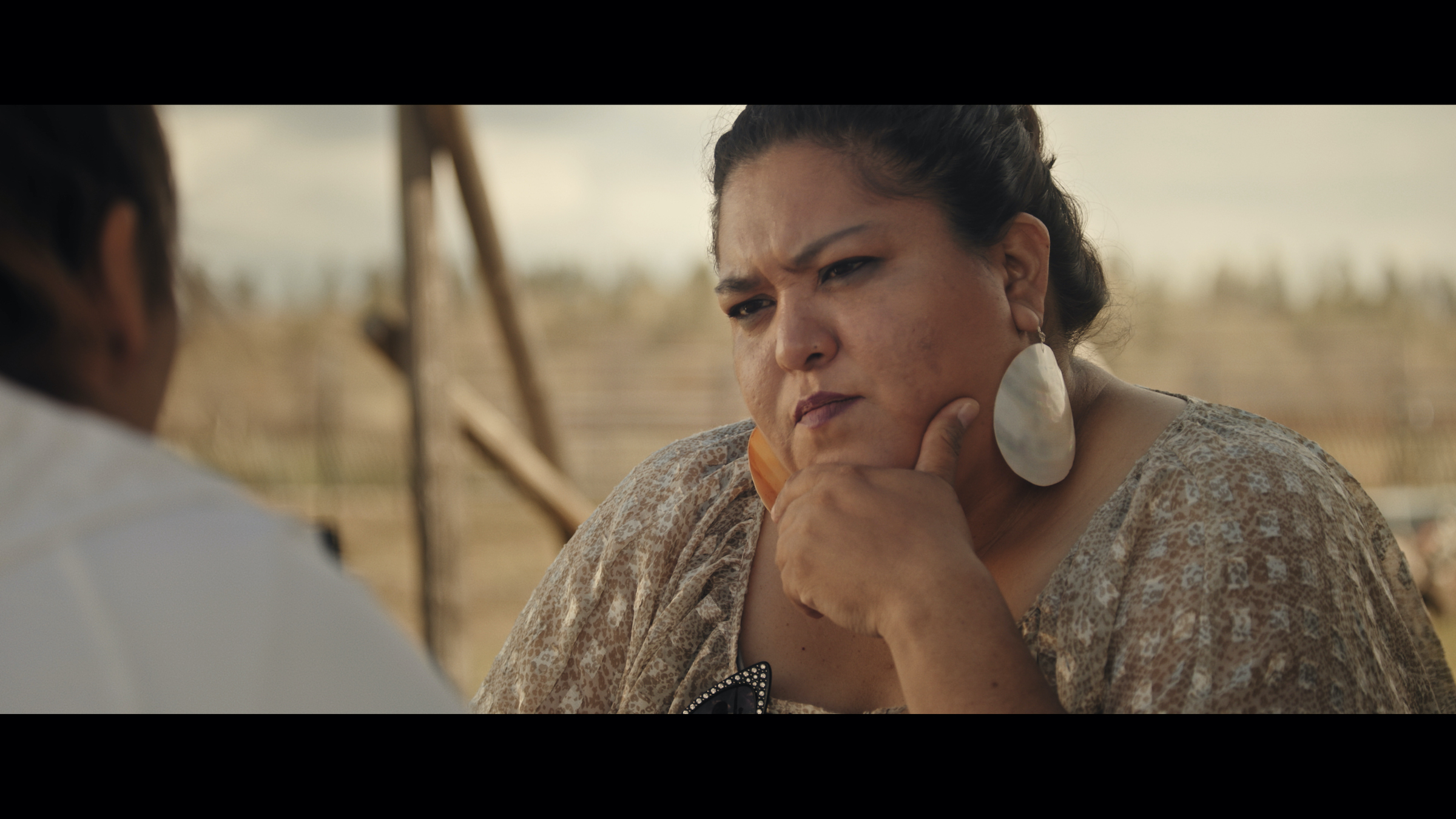 Luella Brien, wearing big dangly shell earrings, looks pensively toward Nakia, sitting just off screen.