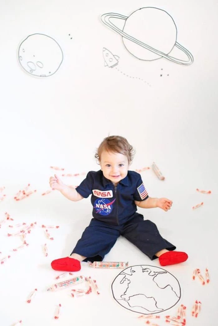 little boy, sitting on the floor, dressed in a nasa onesie, halloween costumes for girls, planets drawn on the wall