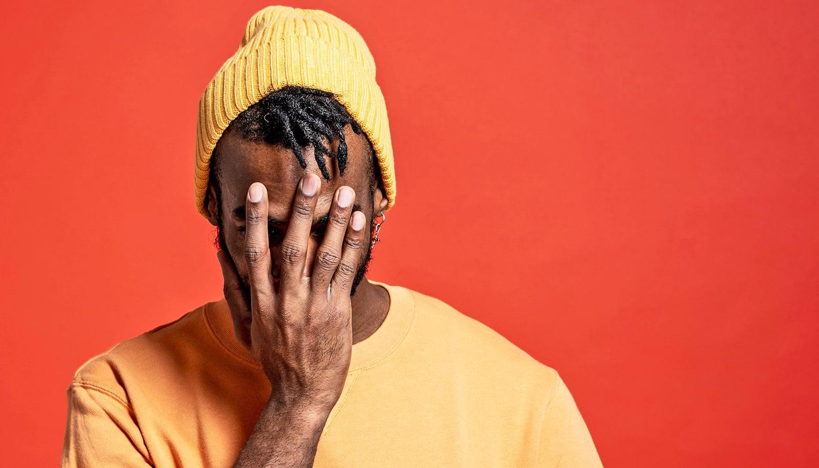 A young man in a yellow beanie and sweatshirt puts his hand over his face.