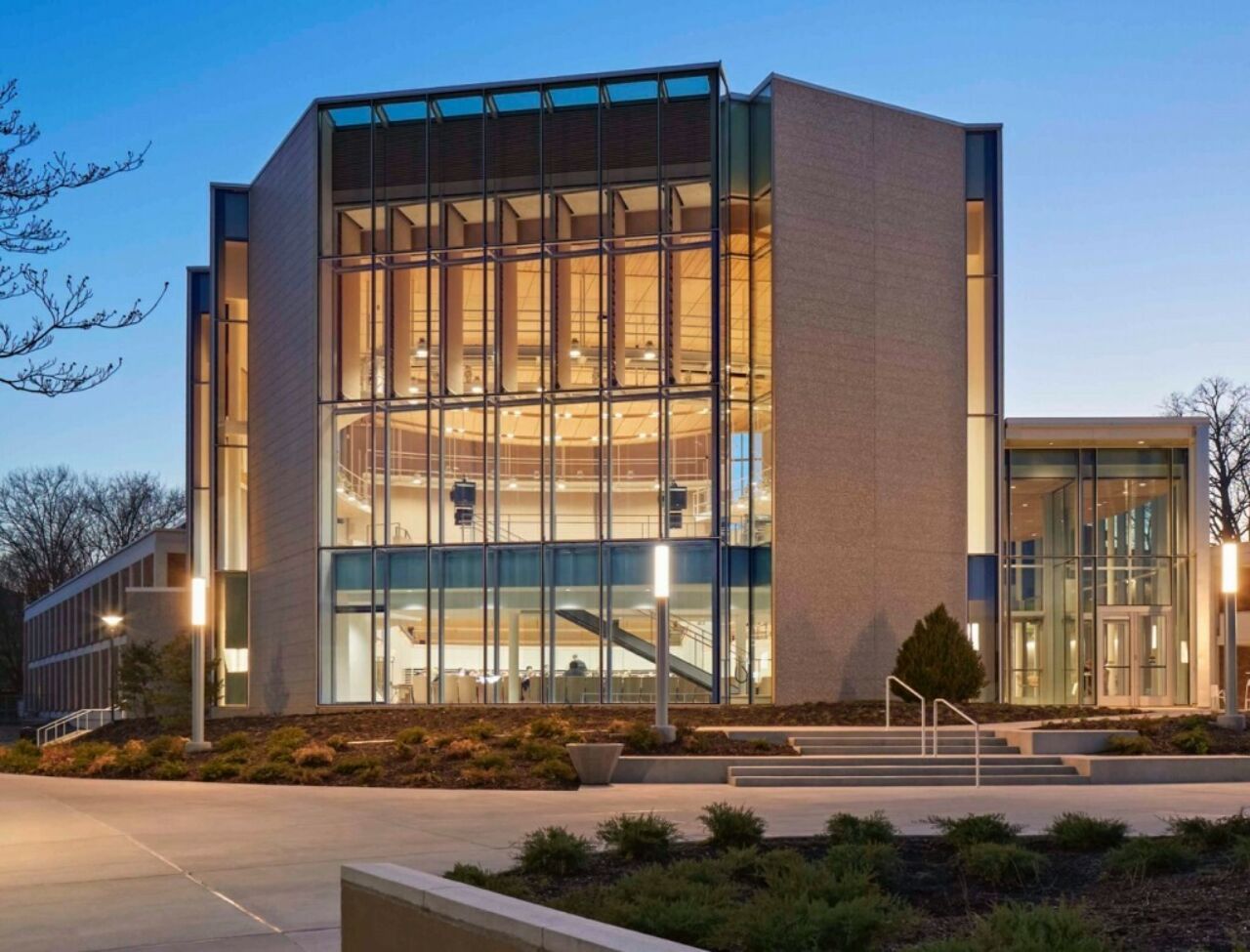 The modern glass facade of Esber Recital Hall illuminating an evening on University Park campus.