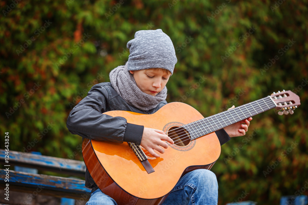 Little boy playing guitar in autumn cold day. He is dressed in a