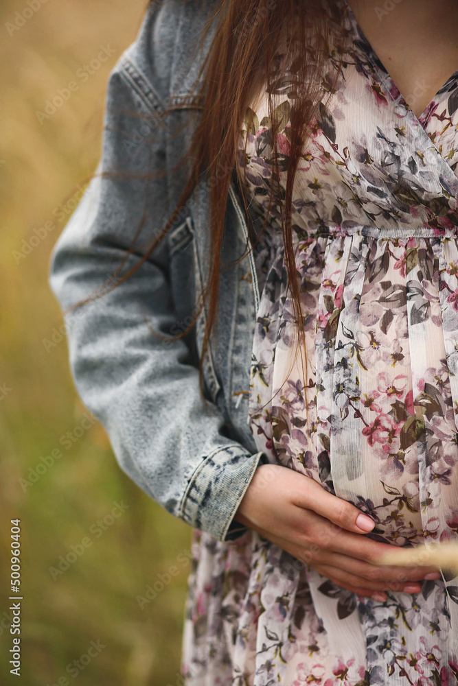 Pregnant woman in beautiful flowery dress and denim jacket touching her ...