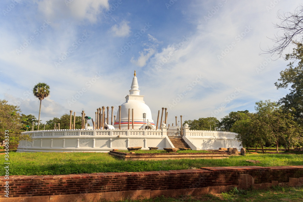 Panoramic view of restored Thuparamaya Dagoba with white bell shaped Stupa, altars, tilted vatadage pillars and Buddhist flags, Anuradhapura, Sri Lanka. Beautiful cloudy sky landscape.