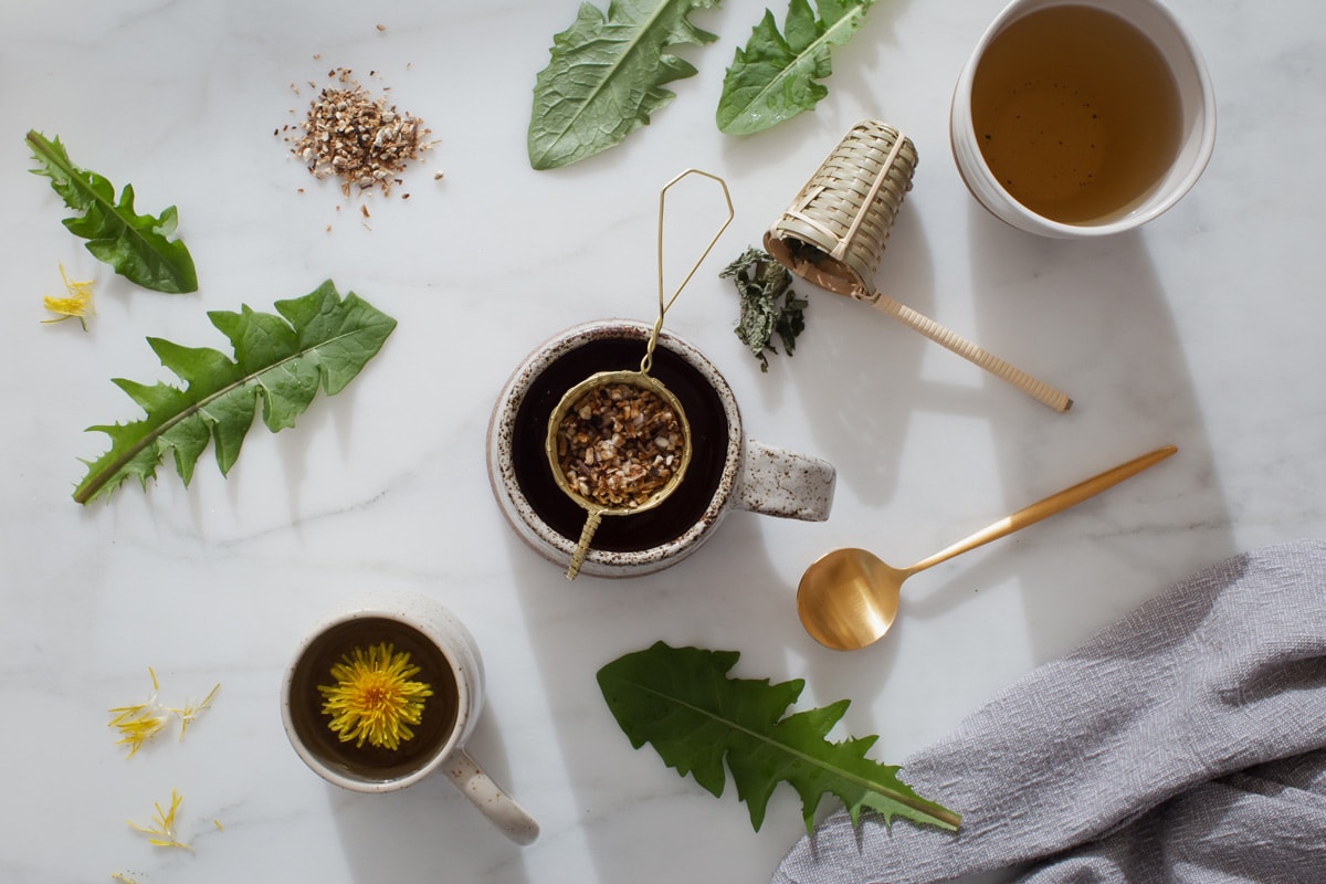 Ceramic mugs holding dandelion flower, leaf and root tea, with various tea strainers, dandelion leaves and flower heads strewn about.