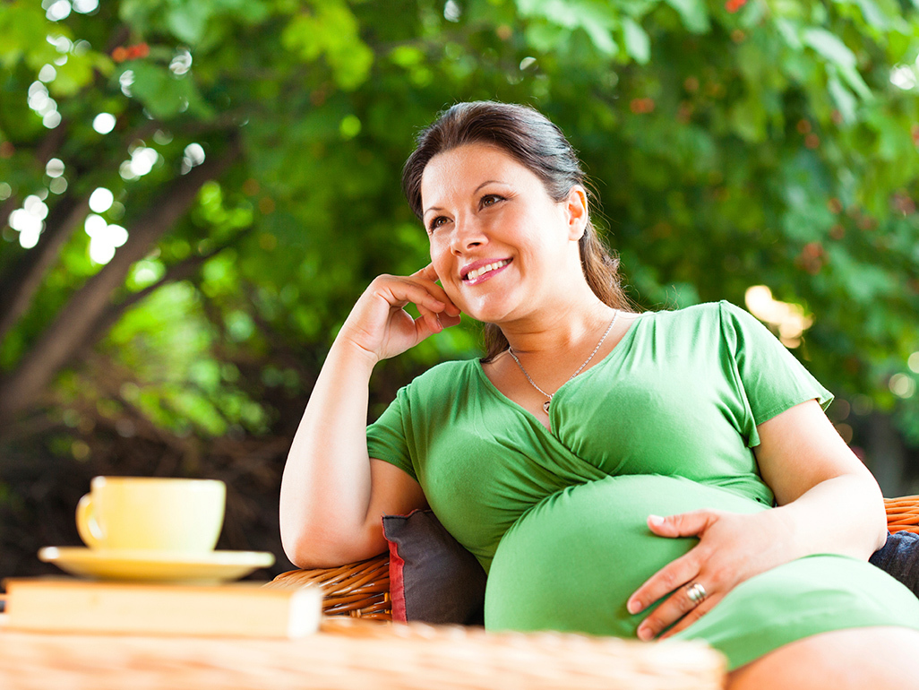 smiling pregnant woman in a green dress, sitting outside with a cup of coffee