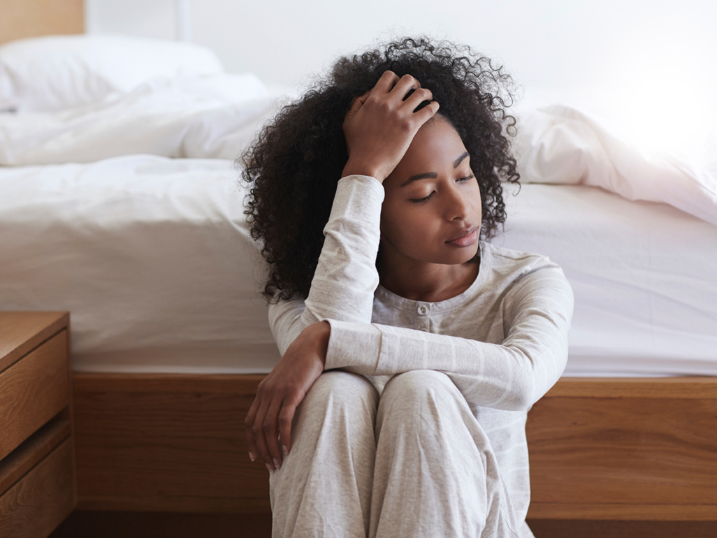 A woman that may be depressed, leaning against her bed, with her hand on her head. 