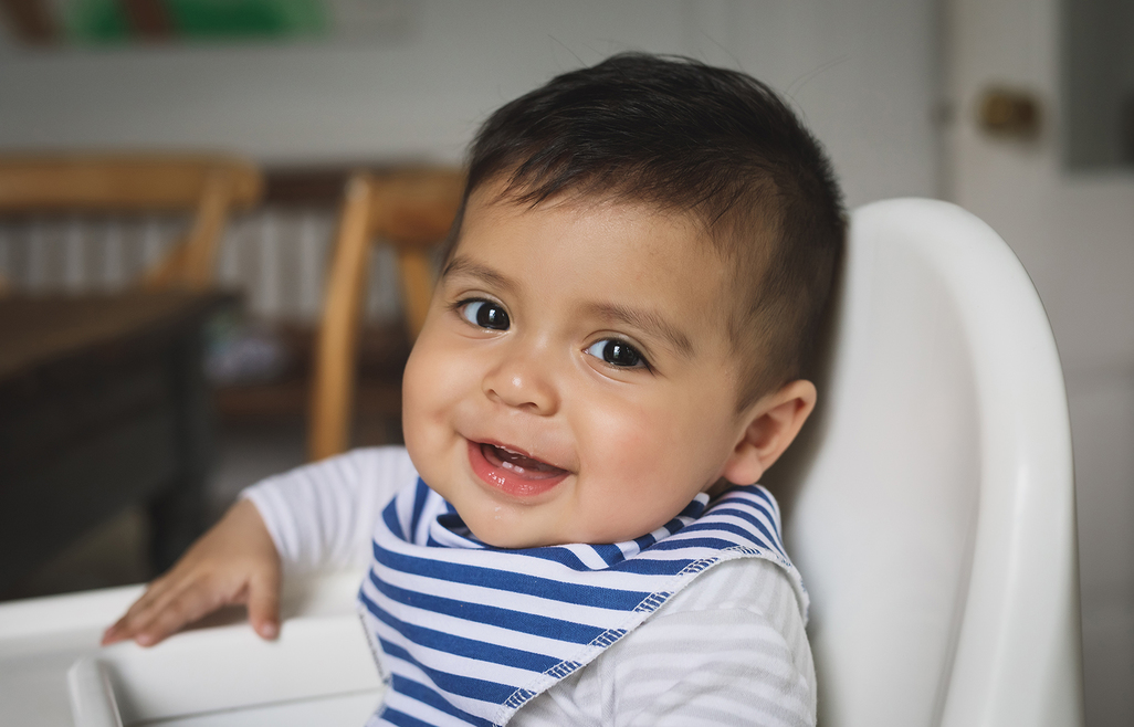 Hispanic baby boy sits in high chair and smiles.