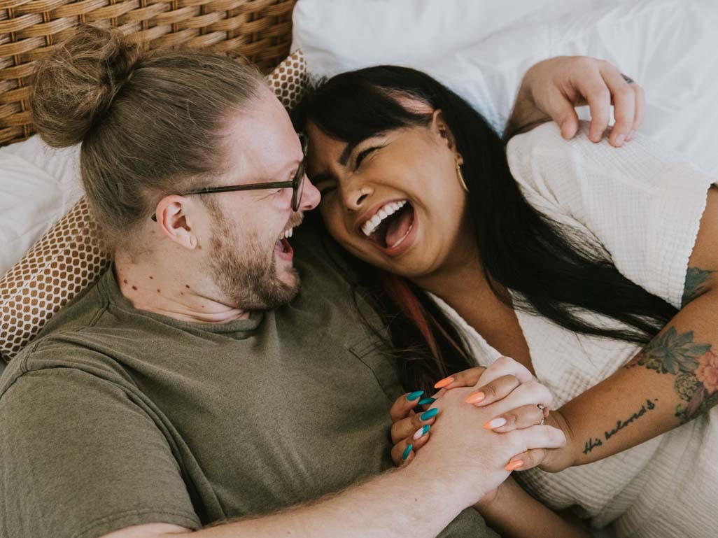 Couple lying in bed holding hands and laughing.