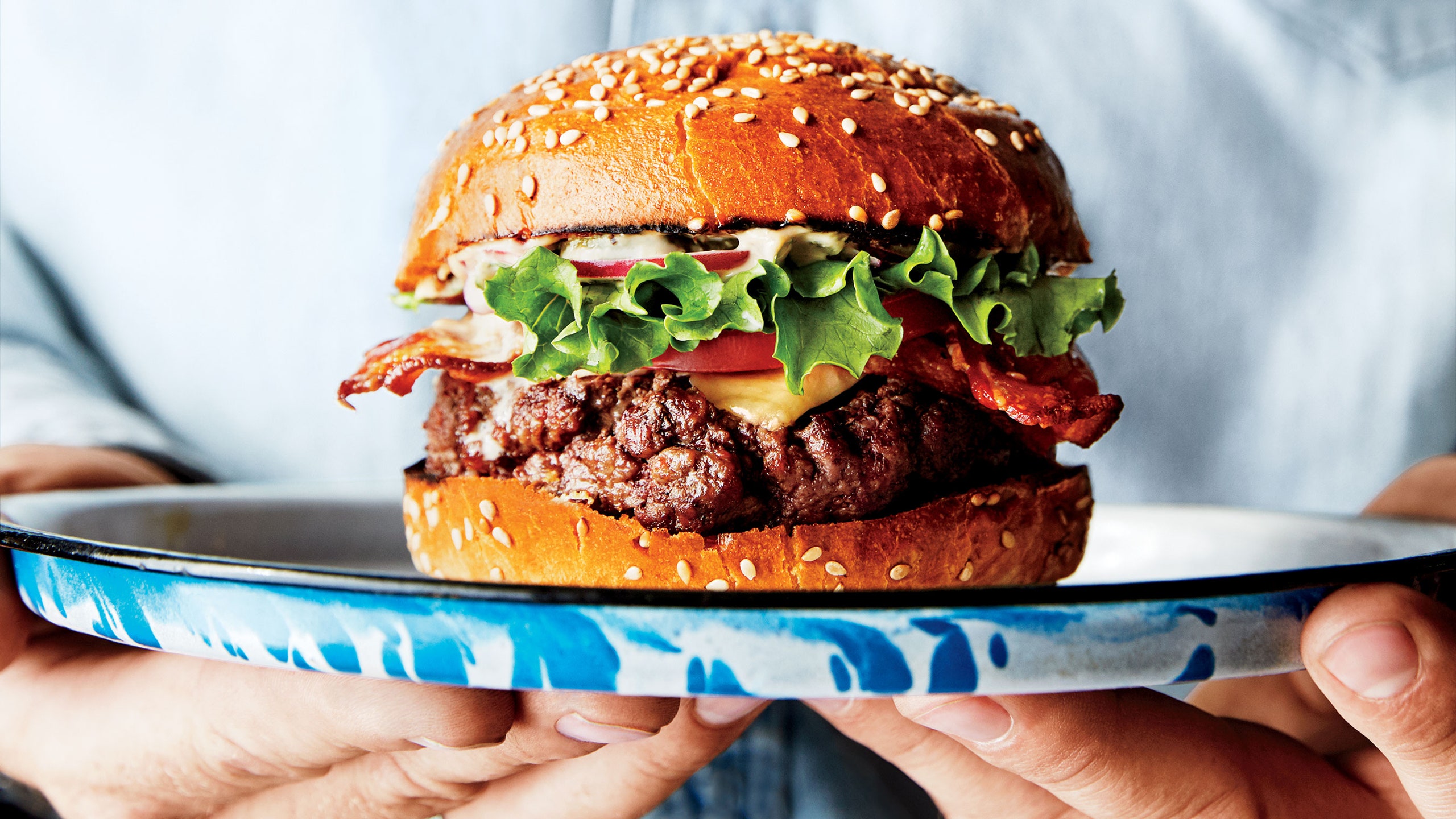 Human hands holding a plate topped with a bacon cheeseburger.