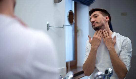 Peson examining their patchy beard in the bathroom mirror