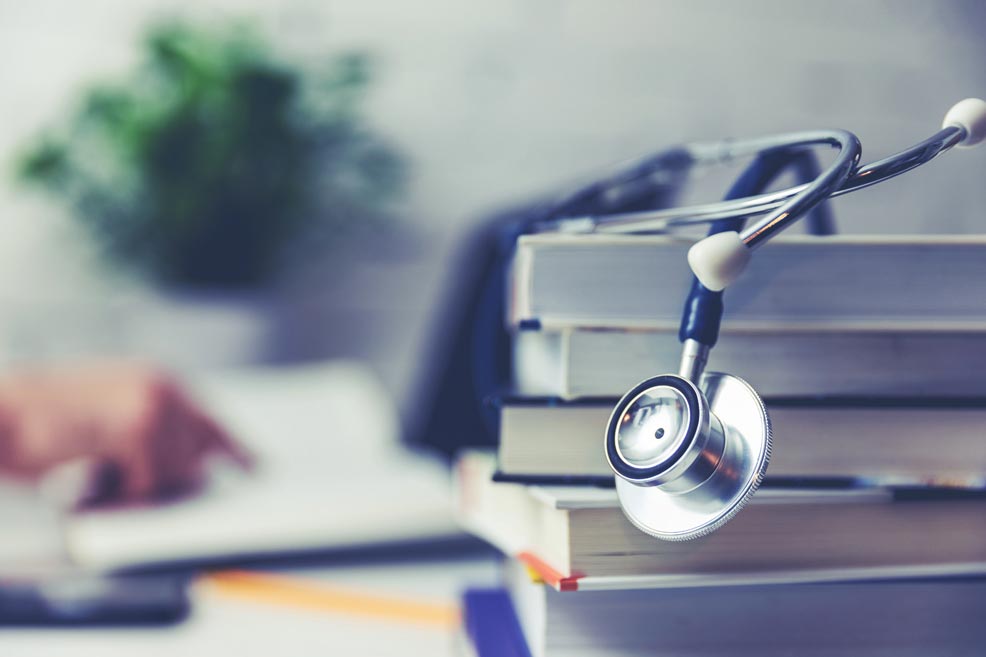 Stack of books with a stethoscope on top in the foreground with a hand pointing into a book in the background